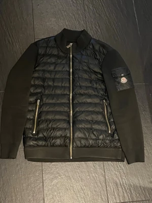 Svart pufferjacka från Moncler - Snygg svart pufferjacka från Moncler storlek L med quiltad front i glansigt material och släta ärmar. Jackan har två dragkedjeförsedda fickor fram och en Moncler-logga på ärmen. Stängs med dragkedja och har en klassisk krage. Perfekt för kalla dagar.