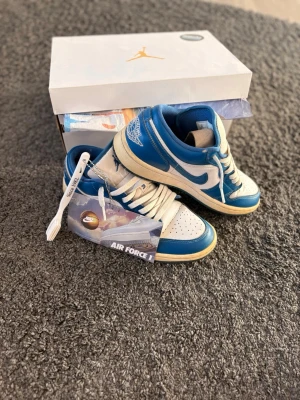 Blåvita Air Jordan 1 Low sneakers - Snygga Air Jordan 1 Low sneakers i blått och vitt med klassisk Nike Swoosh på sidan och Air Jordan-logga på hälen. Ovandelen är i läder och sulan är vit med beige inslag. Perfekta för dig som vill ha en ikonisk och sportig look.