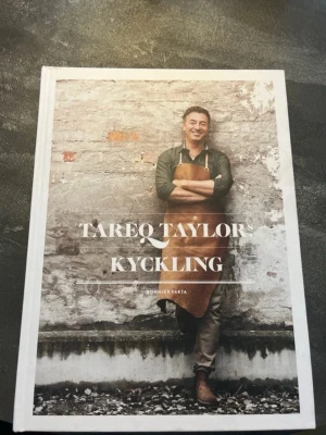 Tareq Taylors Kyckling bok - En snygg kokbok med hårda vita pärmar och ett modernt omslag där Tareq Taylor står mot en tegelvägg. Boken fokuserar på kycklingrecept från hela världen och innehåller inspirerande bilder och tips på hur man lagar kyckling på nya sätt.