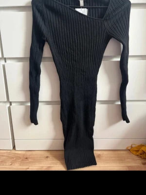 Svart ribbad långärmad klänning H&M - Säljer en svart ribbad klänning från H&M med lång ärm och asymmetrisk halsringning. Klänningen är figurnära och har en stretchig passform som framhäver siluetten. Perfekt för dig som gillar minimalistisk och stilren design.