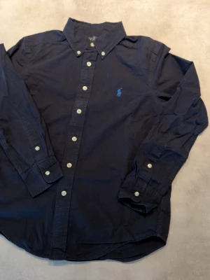 Fräsch skjorta från Ralph Lauren - Riktigt fin marinblå skjorta med ljusblå loga, bara använd ett fåtal gånger och i riktigt gott skick! I barnstorlek M, tveka inte att höra av er kan skickas inom en dag!