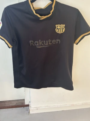 Svart FC Barcelona matchställ - Snyggt svart matchställ från FC Barcelona med guldiga detaljer. Tröjan har klubbmärke och Rakuten-logga på bröstet samt guldfärgade kanter vid hals och ärmar. Shortsen är svarta med klubbmärke i guld. Perfekt för fotbollsträning eller match. Den är äkta och står Messi på ryggen.