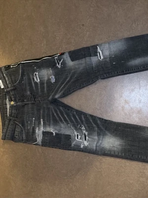 Svarta slitna jeans från Dsquared2 - Säljer ett par svarta jeans från Dsquared2 med slitningar och trasiga detaljer. Jeansen har en tvättad look, fem fickor och dragkedja med Dsquared2-logga. Materialet är denim med stretch för skön passform. Perfekta för en edgy streetstyle.