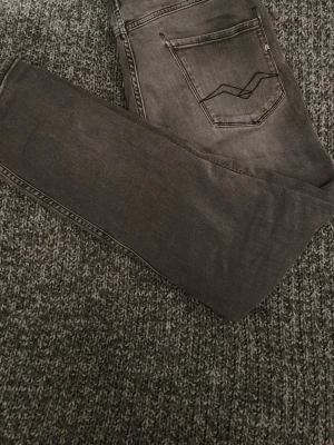 Replay Anbass grå jeans slim fit - Säljer ett par Replay Anbass jeans i snyggt tvättad grå färg. Slim fit-modell med klassiska fem fickor och diskret broderad detalj på bakfickan. Jeansen är tillverkade i mjukt denimtyg som sitter riktigt bra och ger en clean look. Vid minsta fråga är det ba att kontakta mig