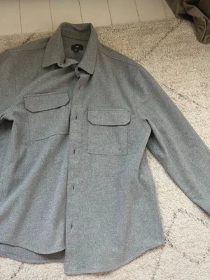 Grå overshirt med fickor - Stilren grå overshirt i mjukt tyg med två stora bröstfickor med lock och knappstängning. Klassisk krage och långa ärmar med knapp vid ärmslut. Perfekt lager-på-lager-plagg för en avslappnad look. Storlek M. Fint skick. 