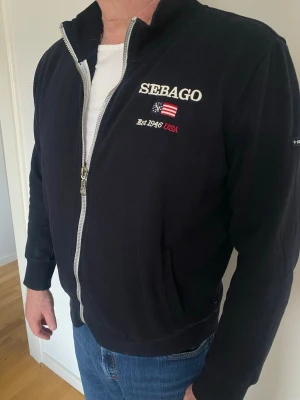 Marinblå ziptröja från Sebago - Snygg marinblå ziptröja från Sebago med broderad logga, amerikansk flagga och texten 'Est 1946 USA' på bröstet. Tröjan har hög krage, dragkedja framtill och ribbade muddar. I jättefint skick.