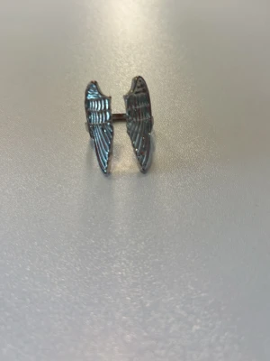 Silverfärgad ring med vingar - Unik ring i silverfärgad metall med två detaljerade vingar som möts framtill. Ringen har en öppen design och vingarna har fina räfflade mönster som ger en cool och stilren look. 