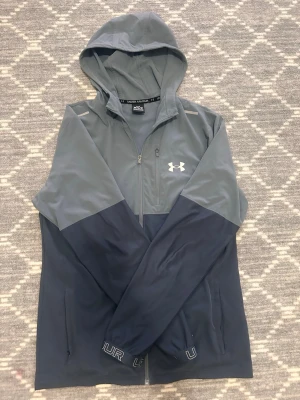 Gråblå vindjacka från Under Armour - Snygg vindjacka från Under Armour i två nyanser av gråblått. Jackan har huva, hel dragkedja framtill, två sidofickor och en bröstficka med dragkedja. Under Armour-logga på bröstet och tryck på ärmsluten. Perfekt för blåsiga dagar. Obs kedjan kan ibland inte funka men det är lätt att fixa.