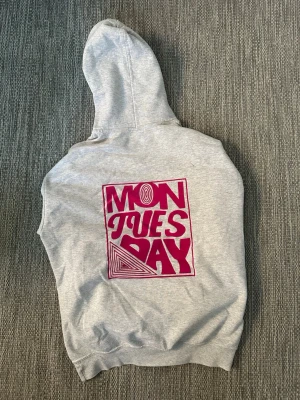 Grå hoodie med tryck från SIZZ - Grå hoodie med stor huva och magficka. På bröstet finns ett litet vinrött SIZZ-tryck och på ryggen ett stort grafiskt tryck i vinrött med texten 'MON TUES DAY'. Tillverkad i mjukt bomullsmaterial.