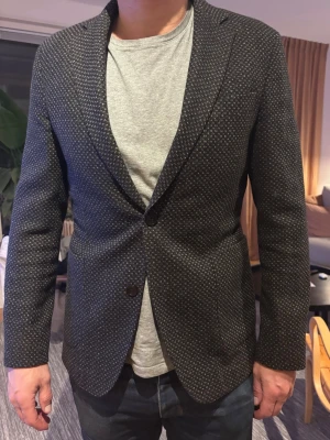 Einar blazer/kavaj från Oscar Jacobsson - Stilren svart kavaj med små vita prickar och klassisk passform. Kavajen har smala slag, enkelknäppning med två knappar och två fickor framtill. Från Oscar Jacobsson modell Einar Blazer nypris ca 4000 kr. Perfekt för en dressad look och passar till både jeans och skjorta. Slim 50 men passar en 48 bättre. Skitsnygg för våren. Skriv för fler frågor!
