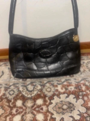 Svart Mulberry handväska  - Snygg svart handväska från Mulberry i krokopräglat läder med axelrem. Väskan har en oval logga framtill och ett guldfärgat emblem vid remmen. Perfekt för dig som gillar klassisk men edgy stil.