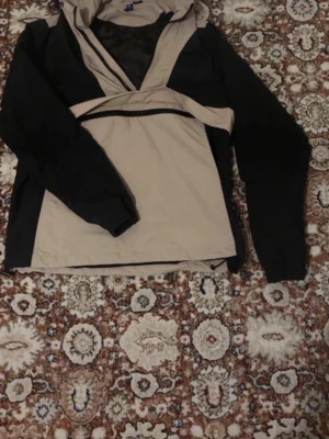 Beige och svart vindjacka med huva - Snygg vindjacka i beige och svart med huva och dragkedja framtill. Jackan har elastiska muddar vid ärmsluten och en stor ficka på magen. Tillverkad i lätt och vindtätt material, perfekt för blåsiga dagar.
