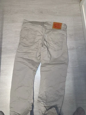 Beiga jeans från Levi's - Säljer ett par beiga jeans från Levi's med klassisk femficksdesign och raka ben. Jeansen har Levi's ikoniska läderpatch bak i midjan och metallknappar. Materialet är bomull och färgen är ljusbeige, perfekt för en avslappnad stil.