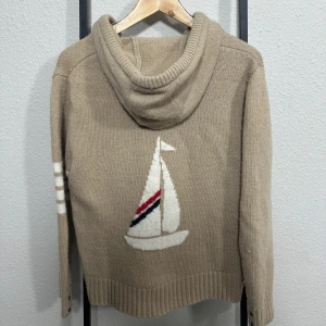 Thom Browné Hoodie ~ Sailboat - 🔹 Beige knitted hoodie in a soft and cozy fabric 🔹 Nautical-inspired design with a large sailboat motif on the front 🔹 Intarsia knit graphic featuring a white sailboat with red and navy stripe accents 🔹 Relaxed silhouette with a slightly cropped hem for a casual look 🔹 Soft hood with a clean, structured knit finish 🔹 Ribbed cuffs and hem for a comfortable and secure fit 🔹 Decorative button detail at the sleeve cuffs 🔹 Sport-inspired white stripes on the sleeve adding subtle contrast