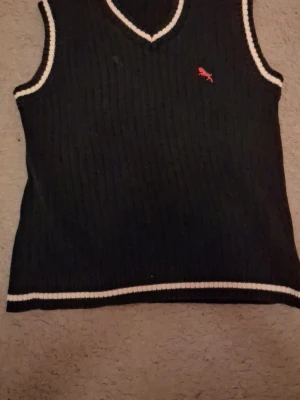 Blå stickad väst med v-ringning - Klassisk svart stickad väst från h&m med v-ringning och vita ribbade kanter. Västen har en liten röd broderad detalj på bröstet och är perfekt för lager på lager. Passar dig som gillar en sportig och stilren look.