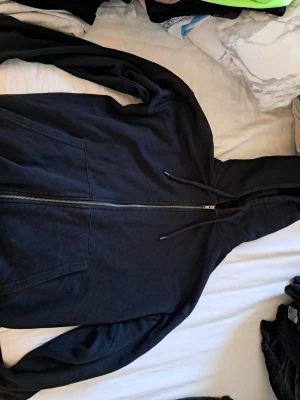 Svart zip hoodie med huva - Klassisk svart hoodie med dragkedja framtill och justerbar huva med snören. Två stora fickor på framsidan och långa ärmar. Perfekt för en avslappnad och stilren look. Materialet känns mjukt och bekvämt, troligen bomullsblandning.