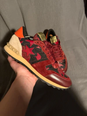 Valentino rockrunner med stjärn design  - Unika sneakers från Valentino i rött och vinrött med svarta och röda stjärnor på sidan. Skorna har detaljer i mocka, läder och textil samt beige och orange häl med nitar. Snygg kontrast mellan färger och material, perfekt för dig som vill sticka ut.