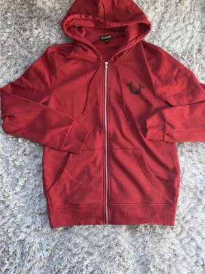Burgundy True Religion Zop Hoodie (S) - Snygg röd hoodie från True Religion med dragkedja framtill och två stora fickor. Klassisk huva med snörning och logga broderad på bröstet. Perfekt för en avslappnad och trendig stil. Den är nästan helt oanvänd och är i toppskick. Den är medium-tunn alltså inte väldigt tjock vilket betyder att den kommer vara perfekt för uppkommande säsonger! Rekommenderas