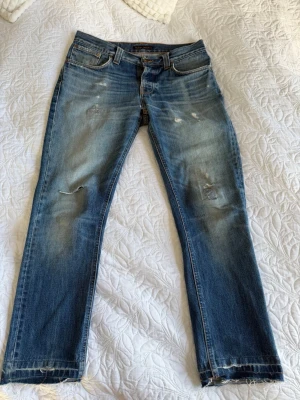 Blå slitna jeans från Nudie Jeans - Säljer ett par blå jeans från Nudie Jeans med slitningar och distressed detaljer på både fram- och baksida. Klassisk femficksmodell med raka ben och ljus tvätt. Snygga kontrastsömmar och patch bak i midjan. Perfekta för en avslappnad streetstil. Jeansen passar bäst för längden 32!! 