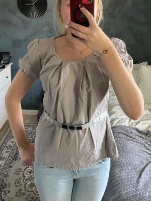 Beige blus med puffärm och bälte - Snygg beige blus med puffiga korta ärmar och rund halsringning. Blusen har ett matchande bälte i midjan med dekorativt spänne och fina veck fram. Perfekt till jeans eller kjol för en trendig look.