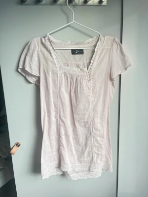 Ljusrosa blus med spetsdetaljer - Söt ljusrosa blus med spetsdetaljer längs halsringning, ärmslut och nederkant. Blusen har korta puffärmar, dekorativa knappar framtill och en feminin, lös passform. Perfekt för dig som gillar romantisk och vintageinspirerad stil. Tröjan är lite genomskinlig så ni är medvetna! 