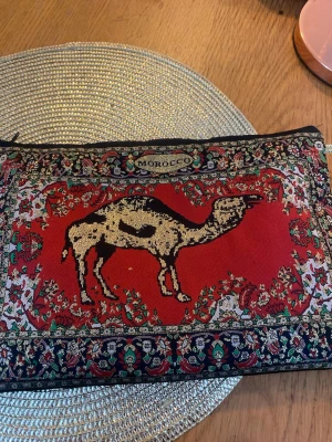 Marockansk handväska med kamelmönster - Unik handväska i tyg med färgstarkt orientaliskt mönster och en stor kamel i mitten. Väskan har dragkedja och en liten ögla för att hålla i. Färgerna är röd, svart, beige och grönt med detaljer i guld och vitt. Perfekt för dig som gillar bohemisk och exotisk stil.