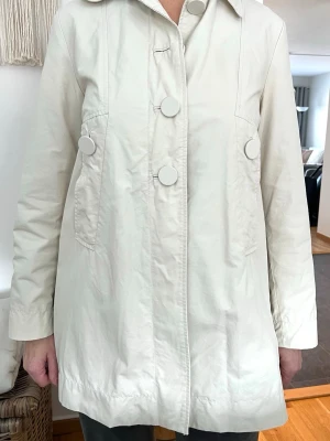 Ljusbeige trenchcoat med stora knappar - Stilren ljusbeige trenchcoat med stora dekorativa knappar framtill och baktill. Jackan har klassisk krage, två fickor och rak passform. Materialet är slätt och lätt, perfekt för vår och höst. Enkel men trendig design som passar till många outfits.