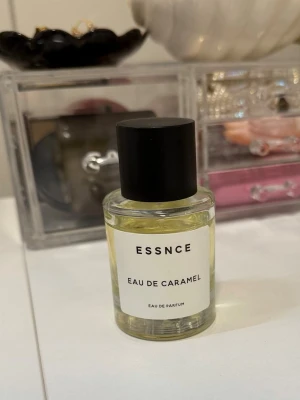 ESSNCE Eau de Caramel parfym - En stilren parfymflaska från ESSNCE med doften Eau de Caramel. Flaskan är cylindrisk i genomskinligt glas med svart plastlock och enkel vit etikett. Innehållet är ljusgult och ger ett modernt och minimalistiskt intryck.
