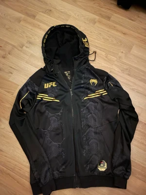 Svart UFC hoodie med guldtryck - Snygg svart hoodie från Venum x UFC med dragkedja, huva och guldfärgade detaljer. Hoodien har tryckta UFC-loggor och mönster i svart och guld på bröstet och ärmarna. Tillverkad i ett sportigt, lätt material som passar perfekt för dig som gillar streetwear och sportig stil.