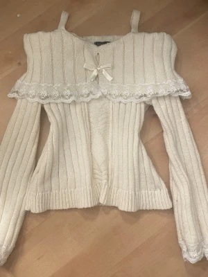 Vit stickad tröja med spets och rosett  - Supersöt vit stickad tröja med ribbad struktur, dekorativ spetsvolang och en liten rosett framtill. Tröjan har långa ärmar, axelband och fina spetsdetaljer med blommönster. Perfekt för dig som gillar gulliga och romantiska plagg.