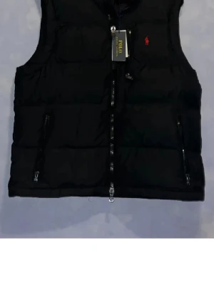 Svart dunväst från Polo Ralph Lauren - Snygg svart dunväst från Polo Ralph Lauren med röd logga på bröstet. Västen har två dragkedjeförsedda fickor fram och quiltad design. Perfekt för lager-på-lager och håller dig varm under kyliga dagar.