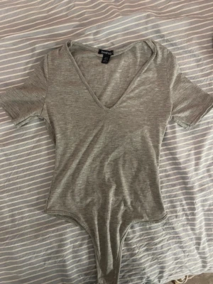Grå body med v-ringning från Boohoo - Snygg grå body från Boohoo med djup v-ringning och korta ärmar. Tillverkad i mjukt stretchigt material som sitter skönt mot kroppen. Perfekt att styla med jeans eller kjol för en clean look.