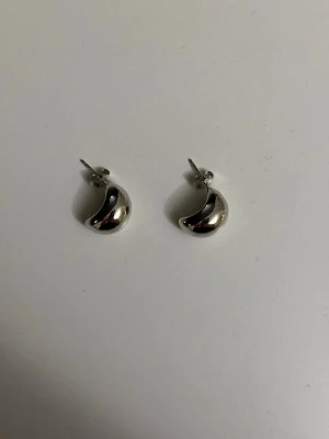 Silvriga chunky örhängen - Säljer ett par silvriga örhängen äkta silver med chunky, böjd form. De har en modern och minimalistisk design som ger ett coolt intryck. Materialet ser ut att vara metall med blank finish. Perfekta för dig som gillar stilrena accessoarer.