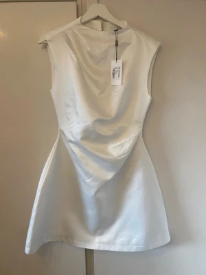 Vit satin kortklänning Oh Polly - Säljer en elegant vit kortklänning från Oh Polly i satin med rak halsringning och ärmlös design. Klänningen har en figurnära passform och en stilren, minimalistisk look som känns både modern och lyxig.