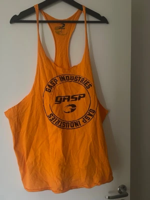 Orange linnetopp från Gasp Industries - Säljer en orange linnetopp från Gasp Industries med stort tryck framtill och brottarrygg. Perfekt för gymmet eller sommarens varma dagar. Luftig passform och tunna axelband ger en sportig vibe.