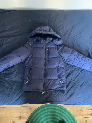 Mörkblå pufferjacka från Polo Ralph Lauren - Snygg mörkblå pufferjacka från Polo Ralph Lauren med huva och dragkedja framtill. Jackan har två stora fickor med dragkedja och en liten röd logga på bröstet. Perfekt för kalla vinterdagar tack vare det tjocka, vadderade materialet.