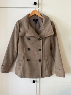 Beige skepparkavaj från H&M - Stilren beige skepparkavaj från H&M med dubbelknäppning och bred krage. Jackan har två snedställda fickor fram och dekorativa knappar bak. Tillverkad i ett mjukt tyg som ger en klassisk look. Perfekt för dig som vill ha en tidlös och snygg jacka. Märkt stl 38 men  liten i storlek, mer stl 36. 