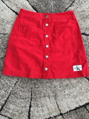 Röd kortkjol från Calvin Klein Jeans - Snygg röd kortkjol från Calvin Klein Jeans med raka linjer och två stora fickor framtill. Kjolen har knappar längs hela framsidan och en liten CK-logga nere till höger. Perfekt för dig som vill sticka ut med färg! Priset är diskuterbart 