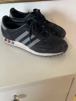 Svarta Adidas LA Trainer sneakers - Klassiska svarta Adidas LA Trainer sneakers med gråa ränder på sidorna och vit sula. Skorna har mesh- och mockadetaljer samt de ikoniska pluggarna i sulan. Lågt skaft och snörning framtill. Perfekta för en sportig och retro look.