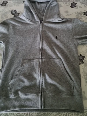 Grå zip hoodie från Ralph Lauren - Snygg grå hoodie med dragkedja från Ralph Lauren. Klassisk design med huva, känguruficka och den ikoniska lilla loggan broderad på bröstet. Tillverkad i mjukt sweatshirtmaterial som är perfekt för chill dagar. Ribbstickade muddar vid ärmslut och nederkant.