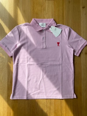 Ljusrosa pikétröja från Ami - Snygg ljusrosa pikétröja från Ami med klassisk krage och två knappar framtill. Tröjan har ett rött broderat hjärta på bröstet och är tillverkad i mjuk bomull. Perfekt för en avslappnad men stilren look.