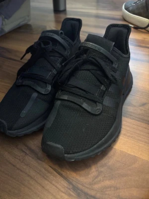 Svarta Adidas sneakers - Säljer ett par helsvarta Adidas sneakers med låg profil och modern design. Skorna har ovandel i mesh för bra andningsförmåga, rejäla svarta snören och detaljer i gummi. Perfekta för dig som gillar stilrena och sportiga skor.