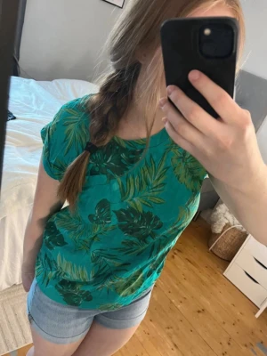 Grön topp med tropiskt mönster - Snygg grön topp med tropiskt bladmotiv i olika gröna nyanser. Modellen är kortärmad och har en avslappnad passform. Perfekt för varma dagar och ger en fräsch sommarkänsla. Tillverkad i mjuk bomull som känns skön mot huden. Finns en liten ficka på bröstet. Säljer även shortsen! 