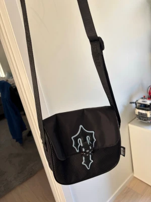 Svart axelväska från Sweet SKTBS - Snygg svart axelväska från Sweet SKTBS med justerbar rem och coolt tryck på locket. Väskan har en praktisk ficka framtill och stängs med spänne. Tillverkad i slitstarkt syntetmaterial, perfekt för att bära det viktigaste.