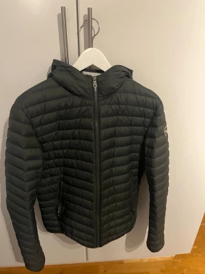 Mörkgrön pufferjacka från Colmar - Snygg mörkgrön pufferjacka från Colmar med huva och dragkedja framtill. Jackan har quiltad design, två sidofickor med dragkedja och Colmar-logga på ärmen. Perfekt för våren och riktigt skön att bära.