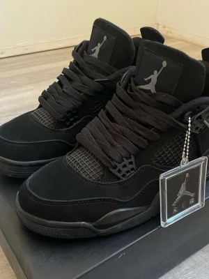 Svarta Air Jordan 4 Retro sneakers - Säljer ett par helsvarta Air Jordan 4 Retro sneakers med ikoniska Jumpman-loggan på plösen. Skorna har meshpaneler, tjocka svarta skosnören och detaljer i mocka och läder. Klassisk siluett med högt skaft och robust yttersula. Perfekta för dig som gillar streetwear.