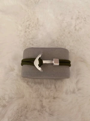 Grönt armband med ankardetalj - Stilrent armband med mörkgrönt, dubbelt snöre och en blank silverfärgad ankare som lås. Armbandet har även fyrkantiga metalldetaljer som ger en modern touch. Perfekt accessoar för dig som gillar marina vibbar och enkel design.