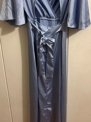 Blå Sommarklänning  - Elegant ljusblå långklänning med omlottliv, vida korta ärmar och ett snyggt knytband i midjan. Klänningen har en mjuk satinliknande yta som ger en lyxig känsla och ett fint fall. Perfekt för dig som vill sticka ut med en stilren look.