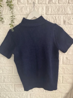 Mörkblå stickad topp med polokrage - Mörkblå stickad topp med ribbade korta ärmar och hög polokrage. Tröjan har även ribbad nederkant och är gjord i ett mjukt material som känns skönt mot huden. Perfekt för lager på lager och en stilren look.