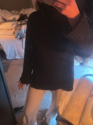 Svart ribbstickad polotröja - Mysig svart ribbstickad polotröja med hög krage och långa ärmar. Tröjan har en avslappnad passform och slitsar i sidorna för extra komfort. Perfekt för lager på lager och enkel att matcha med olika stilar.